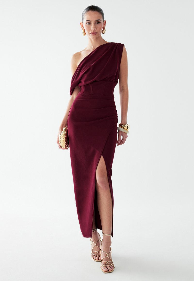 Burgundská, jednoramenná šaty so zhrnutým dekoltom, priliehavým pásom a vysokým rozparkom, vyrobené z hladkej látky, doplnené zlatými sandálmi na opätku.