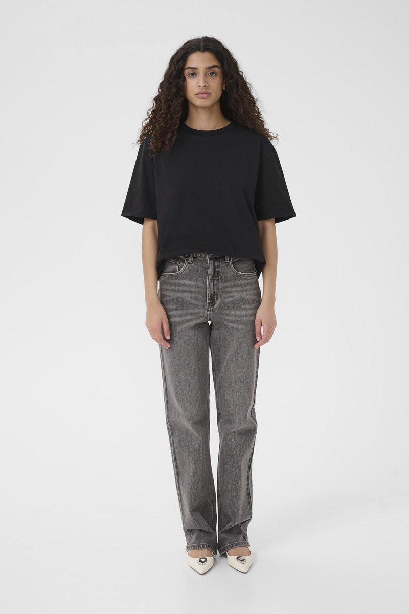 T-shirt en coton noir à manches courtes, associée à un jean en denim gris taille haute avec un léger effet délavé et une coupe droite. Chaussures blanches à bout pointu.