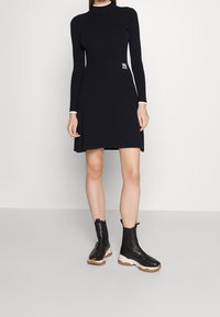 Robe noire côtelée à manches longues et col haut, avec un accent logo blanc. Assortie à des bottines noires à bout et semelle texturée.
