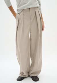 Pantaloni beige plissettati con una vestibilità ampia, realizzati in tessuto leggero. Presentano una vita alta e un design con pieghe frontali. Indossati con scarpe scure.