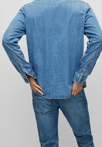 Camisa de denim azul claro con mangas largas, puños abotonados y dobladillo recto. Llevada con unos pantalones de denim azul claro a juego.