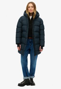 Abrigo acolchado azul marino con capucha, que presenta un diseño acolchado, cremallera frontal y bolsillos laterales. Lleva puesto un suéter de punto negro y jeans azules.