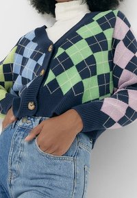 Pletený kardigan se vzorem diamantů v modré, zelené, námořnické a růžové barvě, s dřevěnými knoflíky a žebrovanými manžetami. Nositelské s vysokými džíny.