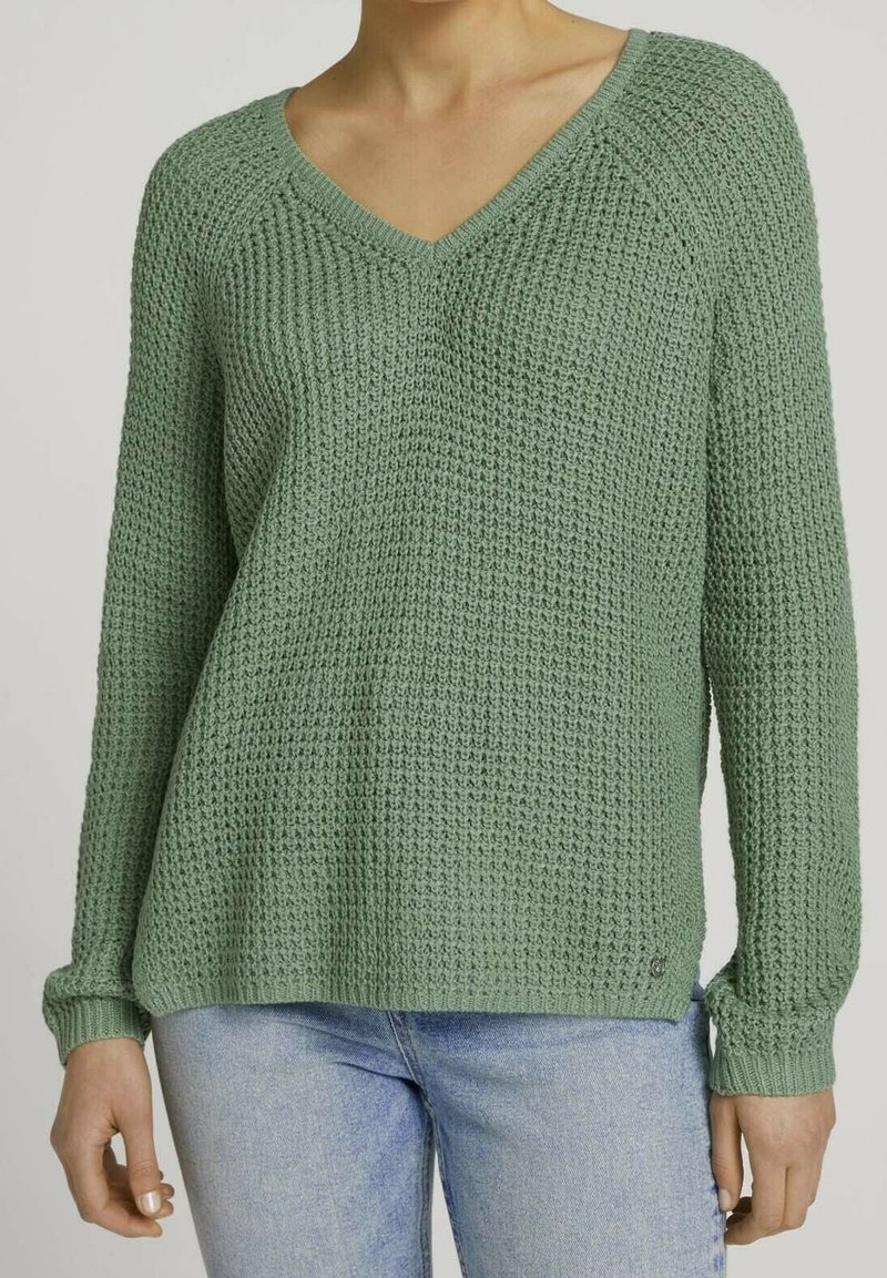 V-ringad tröja med långa ärmar i texturerad grön stickning. Har ribbad fåll och ärmslut, en liten sidoklyfta och en liten logodetalj på nederkanten.