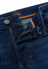 Denim jeans in dark blue with a smooth texture, featuring a visible zipper, orange stitching, and a branded tag inside the waistband.
