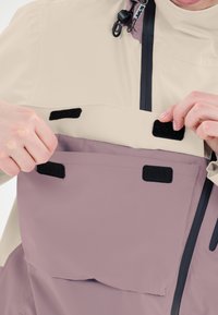 Vattentät jacka med en krämfärgad överdel och lila underdel, med svarta kardborrefästen och en framficka med dragkedja.