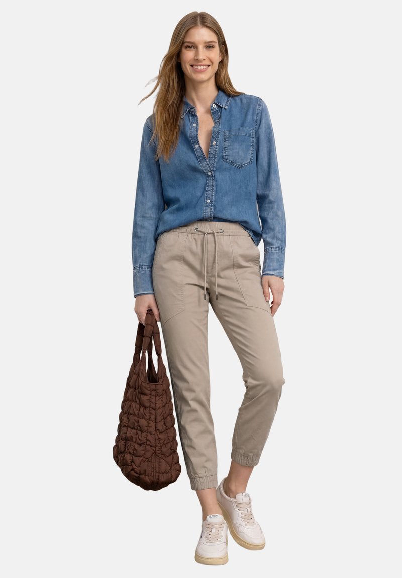 Femme portant une chemise en jean, un jogging beige avec cordon, des baskets blanches, tenant un grand sac matelassé marron, debout devant un fond blanc.