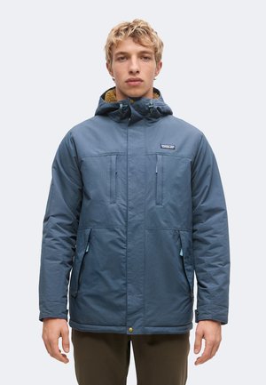 Giovane uomo con capelli biondi corti che indossa una giacca invernale blu con cappuccio e tasche frontali su uno sfondo chiaro e uniforme.