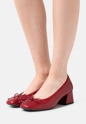 Zapatos de tacón bloque de cuero rojo con puntera redondeada, que presentan un lazo decorativo en el empeine y una superficie texturizada.