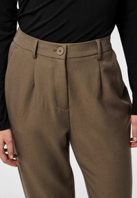 Khakihosen mit glatter Struktur, mit einem einzigen Knopfverschluss, Falten und seitlichen Taschen. Ein schwarzes Langarmoberteil ist teilweise sichtbar.