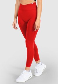 Rote Leggings mit hohem Bund, aus dehnbarem Material. Weiche Textur mit schmalem Schnitt, kombiniert mit weißen Sportschuhen.