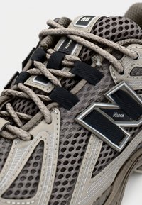 Grå och beige löparskor med meshtopp, plastdetaljer och flätade skosnören. Märkbar "N"-logotyp och ventilationshål finns.