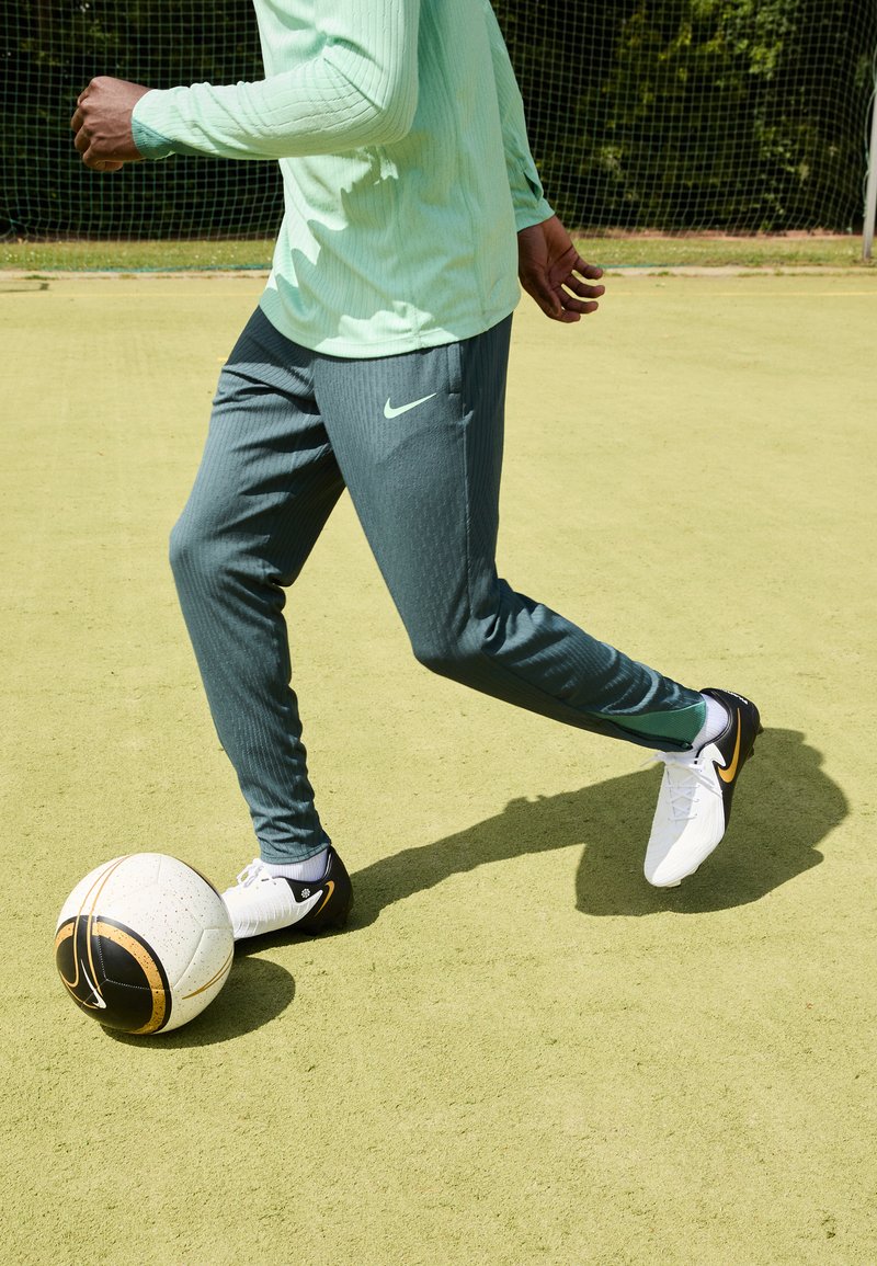 Tenue de sport Nike comprenant un haut à manches longues vert clair et un pantalon rayé vert foncé, associé à des chaussures de football noires et blanches, en train de dribbler un ballon.