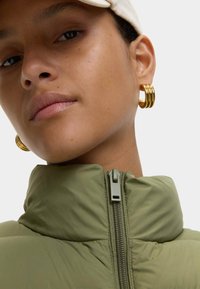 Olijfgroene pufferjack met een hoge kraag en een rits aan de voorkant. Gouden hoopsieraden accentueren het gezicht van het model. Gladde, matte textuur.