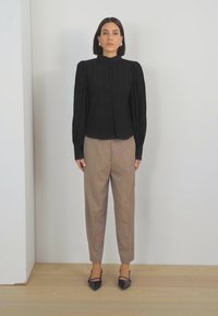 Blouse noire à col montant côtelé avec des manches bouffantes, associée à un pantalon rayé beige clair et des escarpins noirs à bout pointu, debout sur un parquet en bois.