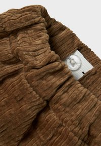 Bruine corduroy stof met een geribbelde textuur en een zichtbare knoopdetail op een wit stoffen label. Zachte en gestructureerde uitstraling.