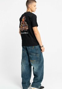 Czarny bawełniany t-shirt z graficzną ilustracją na plecach, zestawiony z luźnymi niebieskimi denimowymi spodniami typu cargo z kieszeniami i detalami szwów.