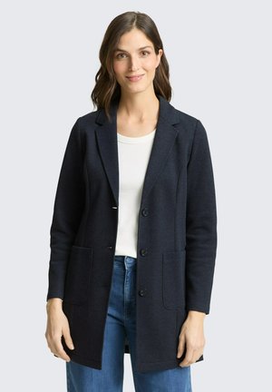 Femme portant un blazer bleu marine foncé sur une chemise blanche et un jean bleu, debout devant un fond gris clair uni.