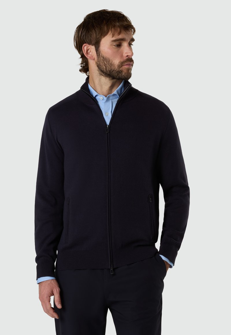 Hombre con barba que lleva una camisa azul claro bajo un cárdigan negro con cremallera, posando con una mano en el bolsillo, mirando hacia la derecha.