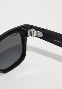 Schwarze rechteckige Sonnenbrille mit dunklen Gläsern, ausgestattet mit einem metallischen Scharnier und silbernen Akzenten. Texturierte Details auf der Oberfläche des Rahmens.