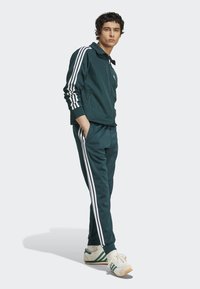 Trainingsanzug aus dunklem grünen Stoff, Zip-Jacke mit weißen Streifen an den Ärmeln, passende Hose und zwei Seitentaschen; getragen mit weißen Turnschuhen.