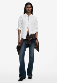 Camicetta bianca ricamata con colletto, abbinata a jeans svasati di colore blu scuro. Giacca di pelle marrone legata in vita. Scarpe nere.