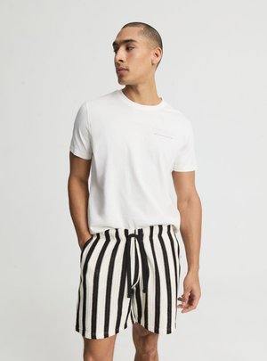 Mężczyzna noszący białą koszulkę z krótkim rękawem oraz czarno-białe pionowo paskowane szorty, z rękami w kieszeniach, patrzący w lewo.