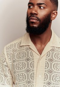 Beżowa koszula z krótkim rękawem, wykonana z geometrycznego, otwartego splotu, z kołnierzykiem i zapięciem na guziki z przodu.