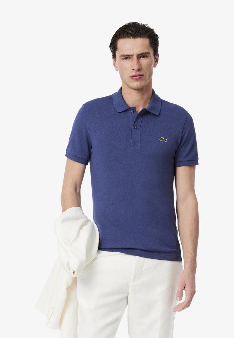 Polo de color azul marino con cuello corto, tapeta de dos botones y un pequeño logotipo verde. Combínalo con pantalones color crema y una chaqueta blanca.
