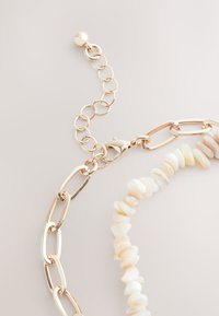 Gold chain necklace featuring a mix of oval links and a shell bead strand in soft beige and white. Includes a clasp and adjustable chain.
