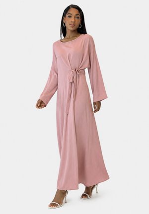 Kvinde iført en lang pink omslagskjole med lange ærmer og hvide sandaletter med hæl, stående mod en ensfarvet lys baggrund.