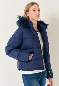 Giacca puffer blu navy con cappuccio in pelliccia sintetica, design trapuntato, due tasche laterali e chiusura frontale con zip. Morbida texture simile alla seta.