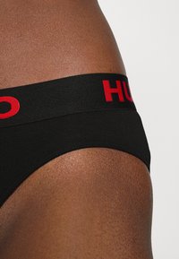 Černé bavlněné boxerky s širokým elastickým pasem, na kterém je červený nápis "HUGO". Tkanina má hladkou strukturu a sedí pod pasem.