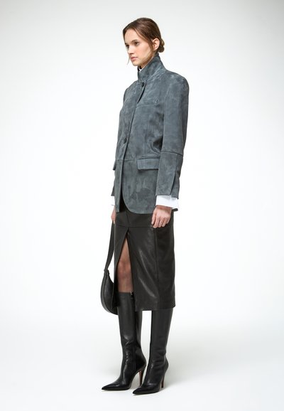 Mujer con blazer gris de cuello alto, falda midi de cuero negro con abertura frontal, botas negras hasta la rodilla y bolso negro en la mano.