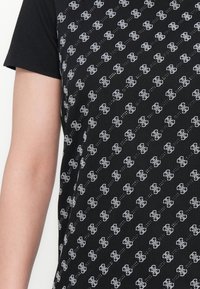 Ingrandimento di una camicia nera a maniche corte con un motivo di logo bianco interconnesso ripetuto e un braccio nudo visibile sul lato sinistro.
