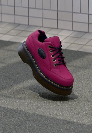 Felroze leren sneaker met zwarte veters en dikke zwarte zool met gele stiksels op een grijze bestratingsachtergrond.