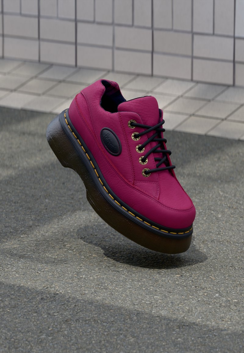 Baskets en cuir rose vif avec lacets noirs et semelle épaisse noire avec surpiqûres jaunes sur un fond pavé gris.
