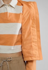 Gehaakte polo met oranje en witte horizontale strepen, gecombineerd met een lichtgewicht, lichtoranje jas met een gaasstructuur en ribbelaccenten.