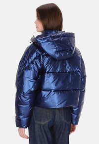 Kék puffert dzseki fényes felülettel, kapucnival és steppelt mintával. Rövidített hosszúság gumírozott mandzsettákkal és szegéllyel. Texturált felület.