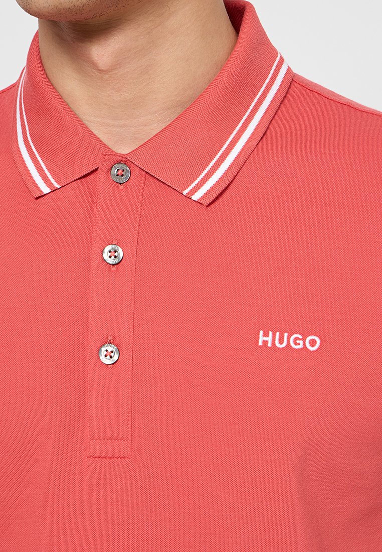 Zbliżenie na osobę noszącą koralowo-czerwoną koszulkę polo z białymi podwójnymi paskami na kołnierzyku oraz haftem "HUGO" na piersi.
