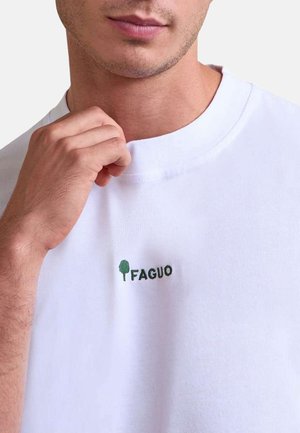 Modèle masculin portant un t-shirt blanc avec un petit logo d'arbre vert et le mot « FAGUO » sur la poitrine, tenant le col.