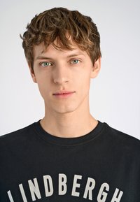 Camiseta de algodón negra con texto "LINDBERGH" en blanco y en una tipografía gruesa. Primer plano de un modelo masculino con cabello castaño claro y ojos azules.