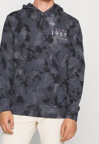 Felpa con cappuccio grigia scura con motivo camouflage. Presenta bottoni sul colletto e una tasca frontale. Stampa "TOMMY 1985 HILFIGER".