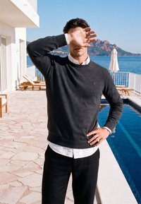 Homme en pull sombre protégeant ses yeux du soleil près d'une piscine, avec des chaises longues et des montagnes en arrière-plan.