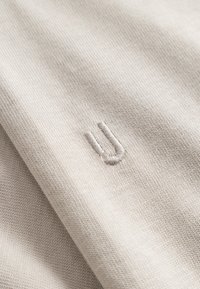 Țesătură texturată de culoare bej deschis cu cusături vizibile. Prezintă un mic logo brodat în culoare asortată, poziționat central pe material.