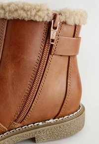 Nahaufnahme eines sandfarbenen Leder-Ankle-Boots mit seitlichem Reißverschluss, Lammfellkragen, gestickter Sohle und strukturiertem Gummi-Außensohle.