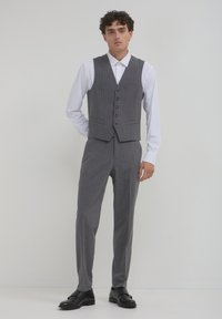 Homme debout portant un gilet à rayures grises et un pantalon assorti, une chemise habillée blanche et des chaussures de ville noires devant un fond clair.