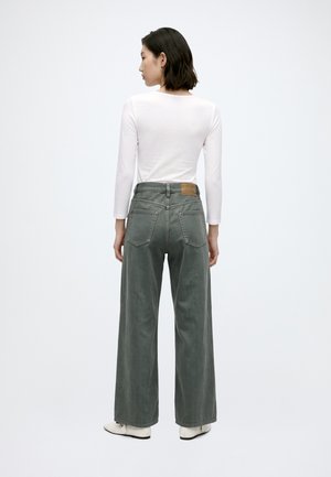 Femme debout, de dos, portant un haut blanc à manches longues, un pantalon gris taille haute à jambes larges et des chaussures blanches sur fond uni.