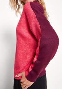 Roter und burgunderfarbener Strickpullover mit einer Mischung aus Texturen, kontrastierenden Ärmelfarben und einem lässigen Schnitt. Weiches Material und runder Kragen.