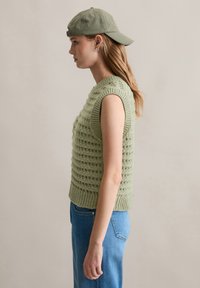 Mouwloze lichtgroene gebreide trui met een gemêleerd patroon; gedragen met een groene pet. Gecombineerd met blauwe high-waisted jeans.